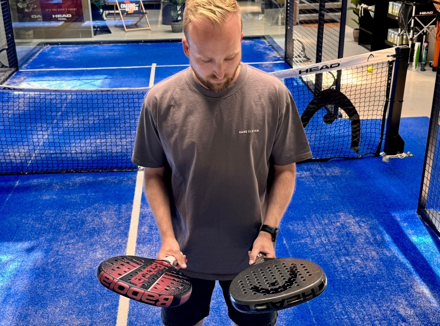 Man håller upp en rosa-svart Babolat padelracket och en svart HEAD padelracket på padelbanan – jämförelse av nybörjarracketar